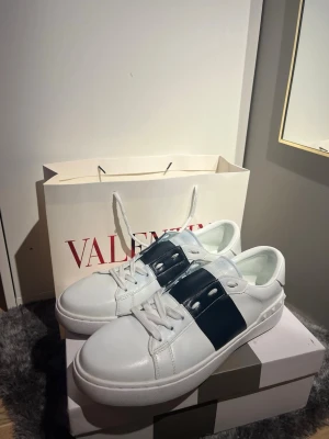 Valentino Garavani Open sneakers vit/blå - Säljer ett par Valentino Garavani Open sneakers i vitt skinn med bred marinblå rand över vristen. Klassisk låg modell med vita snören och coola nitar på hälen. Perfekta för dig som vill ha en clean men ändå statement look. !OBS! Inte orginal snören då dom gick sönder. Men skon är i nyskick