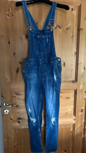 Hängselbyxor i tajt passform och stretchigt jeanstyg - Säljer en blå hängselbyxor i stretchigt jeanstyg. Justerbara hängslen och fickor fram och bak. De har  slitna detaljer på benen och en tajt passform.