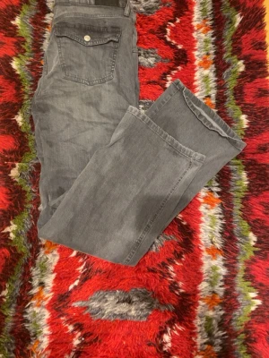 Gråa bootcut jeans med ficklock - Säljer ett par gråa jeans med bootcut passform och snygga ficklock bak med silverfärgade knappar. Jeansen har klassisk femficksdesign och raka ben med lite vidare avslut. Perfekta för dig som gillar en avslappnad men ändå stilren look.