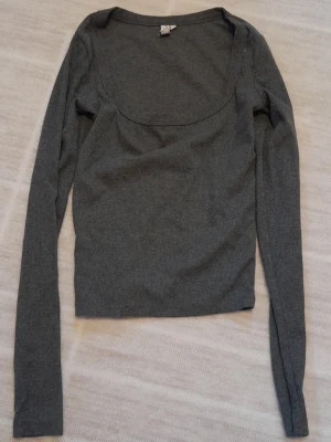 Mörkgrå ribbad topp  - Säljer en mörkgrå ribbad långärmad topp från Nelly i storlek XS. Toppen har en djup, rundad urringning och är tillverkad i mjukt stretchigt material som sitter snyggt på kroppen. Perfekt att styla med jeans eller kjol. !!endast swish +frakt !!