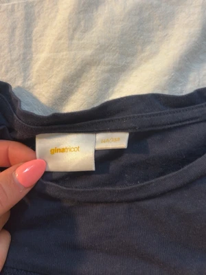 Mörkblå topp med volang från Gina Tricot - Supersöt mörkblå topp från Gina Tricot i storlek 146/152. Toppen har korta ärmar, smockad struktur över hela framsidan och en volangkant längst ner. Tillverkad i mjuk bomull som känns skön mot huden. Perfekt för dig som gillar en enkel men ändå detaljerad look.
