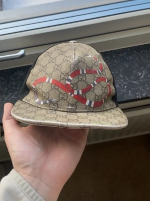 Gucci keps med orm och GG-mönster - Säljer en snygg Gucci keps med klassiskt GG-mönster i beige och svart mesh baktill. Framtill pryds kepsen av en röd och vit orm och justerbart band bak med gröna och röda ränder. Perfekt accessoar för dig som vill sticka ut.
