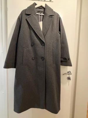 Grå dubbelknäppt kappa från Zara - Säljer en grå, lång och oversized kappa från Zara i ullblandning. Klassisk dubbelknäppt design med stora slag, två fickor framtill och dekorativ knappdetalj vid ärmslut. Perfekt för dig som vill ha en stilren och tidlös look.