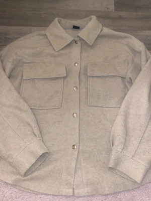 Beige overshirt med fickor - Säljer en beige overshirt med två stora bröstfickor och tryckknappar framtill. Jackan har klassisk krage och är tillverkad i ett mjukt material som påminner om ull. Perfekt för lager på lager och enkel att styla till olika outfits.