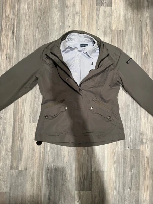 Grå vindjacka från Magliano - Säljer en grå vindjacka från Magliano i storlek M. Jackan har flera fickor med dragkedja och knappar, hög krage och justerbar nederkant. Perfekt för dig som vill ha en stilren och funktionell jacka till vardags. Klassisk passform och snygga detaljer.