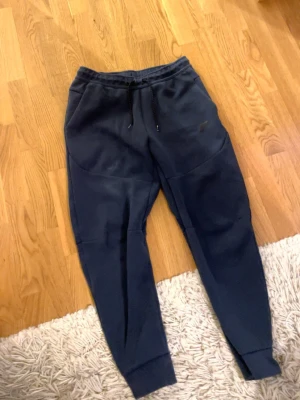Mörkblå mjukisbyxor från Nike - Säljer ett par mörkblå mjukisbyxor från Nike med broderad logga på benet. Byxorna har dragsko i midjan, ribbade muddar vid bensluten och är gjorda i mjukt bomullsmaterial. Perfekta för chill eller träning.