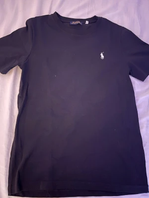 Svart t-shirt från Polo Ralph Lauren - Klassisk svart t-shirt från Polo Ralph Lauren med den ikoniska vita logon broderad på bröstet. T-shirten har rund hals och korta ärmar, tillverkad i mjuk bomull för skön känsla. Enkel och stilren design som funkar till allt.