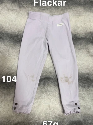 Ljuslila leggings med knappar - Säljer ett par ljuslila leggings från Newbie. De har dekorativa knappar och liten volangdetalj vid bensluten. Perfekta för dig som gillar bekväma och söta byxor till vardags.🌸 Fläckar. Storlek 104