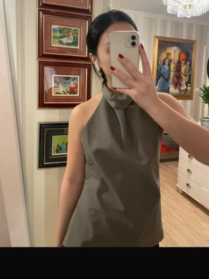 Grå halterneck topp från Zara - Säljer en stilren grå/grön halterneck topp från Zara i storlek S. Toppen har hög hals och är ärmlös med öppen rygg. Är helt ny!