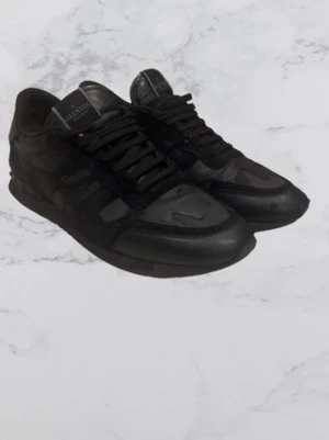 Valentino Garavani svarta sneakers - Säljer ett par svarta Valentino Garavani sneakers med coolt kamouflagemönster och nitar på hälen. Skorna har snörning, rund tå och är tillverkade i en mix av skinn och textil. Perfekta för dig som vill ha en edgy och exklusiv look.