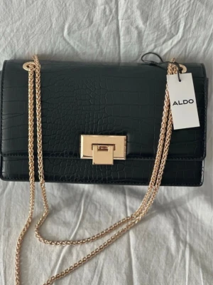 Svart axelväska från ALDO med kedja - Snygg svart axelväska från ALDO i croco-mönstrat syntetmaterial. Väskan har guldfärgad kedjerem och ett stilrent guldfärgat lås framtill. Perfekt storlek för det viktigaste och ger en lyxig vibe till din outfit.