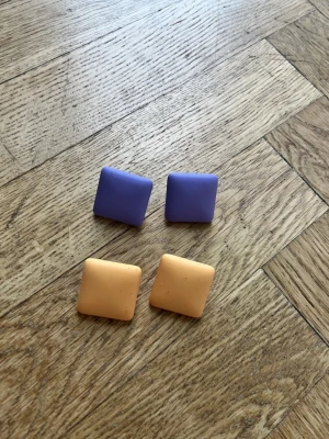 Fyrkantiga lila och orange örhängen - Två par örhängen i cool fyrkantig form, ett par i lila och ett par i orange. De är ca 3x3 cm stora och har metallstift med plastbaksida. Perfekta för dig som vill addera färg till din look.