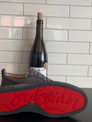 Gråa Louboutin sneakers med röd sula - Snygga grå sneakers från Louboutin med ikonisk röd sula och diskret snörning. Skorna är i mocka och har en stilren, rund tå. Perfekta för dig som vill sticka ut med en lyxig touch och klassisk design. Pris kan diskuteras vid snabb affär 