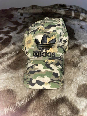 Camouflagekeps med blommor från Adidas - Säljer en keps från Adidas med camouflagemönster i grönt, beige och svart, dekorerad med stora gula blommor. Kepsen har broderad Adidas-logga framtill och är tillverkad i bomull. Perfekt för dig som vill sticka ut med en unik accessoar.