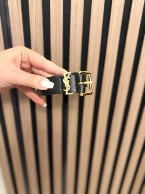 Svart bälte från Yves Saint Laurent - Säljer ett svart bälte från Yves Saint Laurent med guldfärgat YSL-spänne. Bältet är i skinn och har en klassisk, stilren design med exklusiv känsla. Perfekt accessoar för att lyfta vilken outfit som helst.