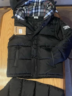 Svart pufferjacka från Burberry *limited stock* - Säljer en svart pufferjacka från Burberry med rutigt foder i huvan. Jackan har dragkedja och knappar framtill, två sidofickor med dragkedja och Burberry-logga på ärmen. Perfekt för kalla dagar och riktigt snygg streetstil.