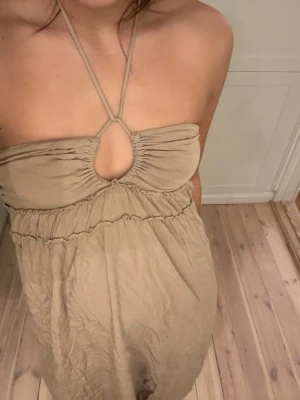 Beige halterneck långklänning - Säljer en beige långklänning med halterneck och snygg cut-out framtill. Klänningen har en luftig passform och smock i ryggen för extra komfort. Perfekt för varma dagar och enkel att styla med accessoarer.