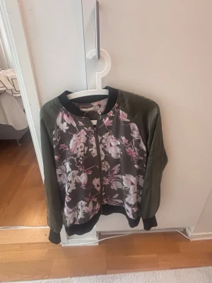 Blommig bomberjacka från Fransa - Snygg bomberjacka från Fransa i militärgrönt med blommigt mönster i rosa och vitt på både fram- och baksida. Jackan har svarta muddar, dragkedja framtill och är tillverkad i ett glansigt polyestermaterial. Perfekt för dig som vill sticka ut lite extra.