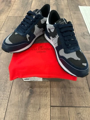 Valentino Garavani Rockrunner sneakers - Snygga Valentino Garavani Rockrunner sneakers i marinblått, svart och vitt med camouflagemönster. Skorna har ovandel i mocka och mesh, svarta snören och en vit sula med svarta detaljer. Perfekt för dig som vill ha en unik och trendig look.