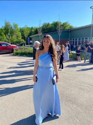 Ljusblå balklänning med oneshoulder - Säljer en elegant ljusblå långklänning i satinliknande material. Klänningen har oneshoulder-design och en mjukt draperad överdel som ger en snygg siluett. Perfekt för bal eller student. Enkel och stilren look som känns både modern och klassisk. Använd 1 gång!