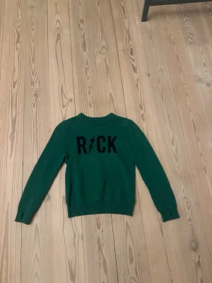 Grön kashmirtröja Zadig & Voltaire 10år - Mörkgrön stickad tröja från Zadig & Voltaire i mjuk kashmir. Framsidan har svart text med 'ROCK' och en blixtdetalj. Klassisk rund halsringning och långa ärmar med ribbade muddar. Perfekt för dig som gillar edgy och stilren design.10år