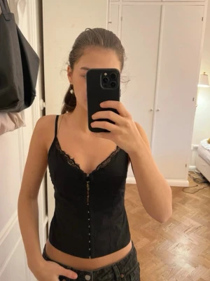 Svart spetsdetaljerad korsett - Säljer en svart korsett med tunna axelband och snygga spetsdetaljer längs v-ringningen. Toppen har knäppning framtill med små krokar och är figurnära i modellen