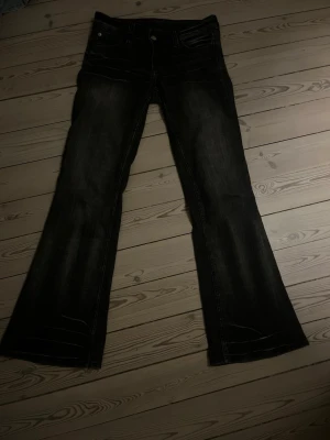 Mörka bootcut jeans med vintagekänsla - Säljer ett par mörkgrå bootcut jeans med snygg tvätt och slitningar. Jeansen har klassisk femficksdesign, låg midja och ett coolt patch på bakfickan. Perfekta för dig som gillar retrovibbar och vill ha ett par jeans som sticker ut. Storlek 28/30 och passar perfekt för mig som är 166 cm lång.
