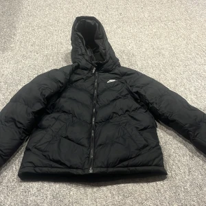 Svart pufferjacka från Nike med huva - Säljer en svart pufferjacka från Nike med huva och dragkedja framtill. Jackan har en clean look med diskret Nike-logga på bröstet och är perfekt för kalla dagar. Tillverkad i syntetmaterial och har långa ärmar för extra värme. Jackan är från 158cm till 170cm alltså barnstorlek XL
