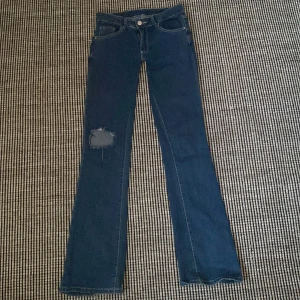 Blå bootcut jeans med slitning - Snygga blå jeans med bootcut och super lowwaist passform och cool slitning på ena knät. Byxorna har markerade bakfickor med lock och knappdetaljer. Klassisk femficksmodell i jeansmaterial, perfekta för en avslappnad och trendig look.