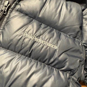 Svart dunjacka Peak Performance - Säljer en svart dunjacka från Peak Performance med huva och dragkedja. Jackan är quiltad med breda kanaler och har diskret logga broderad på bröstet. Perfekt för kalla dagar och har en stilren look som funkar till det mesta. Storlek 160. 