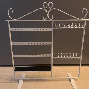 Vit smyckesställning - Snygg vit smyckesställning i metall med hjärtformad detalj upptill. Flera krokar och hål för halsband och örhängen och en svart hylla längst ner för ringar. 