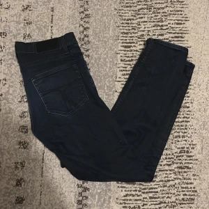 Svarta jeans Pistolero 30/32 - Svarta jeans från Tiger of Sweden i modellen Pistolero, med klassisk femficksdesign och guldfärgade nitar. Jeansen har en rak passform och är tillverkade i ett slitstarkt jeanstyg. Perfekt för dig som gillar stilrena och tidlösa jeans. Jeansen har både en svart och en mörkblå färg som matchar och passar in jätte bra för en unik och snygg look. Priset kan diskuteras för en snabb affär 👍