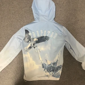 Trapstar windbreaker - Säljer en sjukt snygg ljusblå vindjacka från Trapstar med stort örntryck och logga på ryggen. Jackan har huva, dragkedja framtill och två fickor med dragkedja. Perfekt för dig som vill sticka ut med streetwear-vibe.