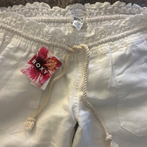 Vita linnebyxor från Roxy med snörning - Snygga vita linnebyxor från Roxy med elastisk midja och grov snörning framtill. Byxorna har raka ben, sidfickor och bakficka med broderad logga. Perfekta för en chill och avslappnad stil under varma dagar. Helt oanvända!