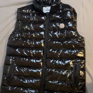 Svart dunväst från Moncler - Snygg svart dunväst från Moncler med glansig finish och klassisk logga på bröstet. Västen har dragkedja framtill, två fickor med dragkedja och quiltad design. Perfekt för lager-på-lager och ger en cool streetstyle-vibe.