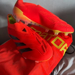 Adidas Predator röda fotbollsskor - Säljer ett par Adidas Predator fotbollsskor i en riktigt fet röd färg med svarta detaljer och transparent sula med gula inslag. Skorna har en strumpliknande passform och är tillverkade i syntetmaterial, perfekta för dig som vill sticka ut på planen. I nyskick endast använda två gånger storlek 39 1/3
