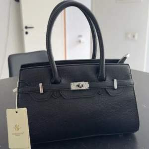 Svart handväska  - Stilren svart handväska i skinnimitation. Väskan har två rejäla handtag, silverfärgade metalldetaljer och medföljande axelrem. Invändigt finns flera fack och ljusbeige foder. Pris kan diskuteras.