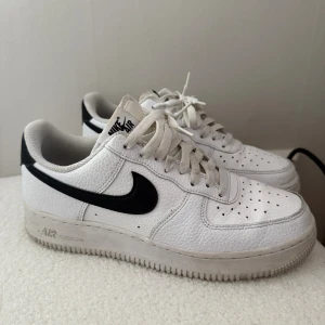 Nike Air Force 1  - Nike Air Force 1 sneakers i vitt skinn med svarta detaljer och klassisk svart swoosh. Lågt skaft, perforerad tå och snörning framtill. Tjock, platt sula och broderad Nike Air-logga på hälen. Perfekta för dig som gillar en clean och tidlös streetstil. I bra skick.