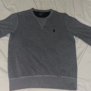 Äkta Grå sweatshirt från Polo Ralph Lauren - Klassisk grå sweatshirt från Polo Ralph Lauren med rund halsringning och broderad marinblå logga på bröstet. Tröjan har långa ärmar och ribbade muddar vid ärmslut och nederkant. Perfekt för en chill och stilren look.