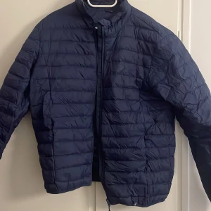 Mörkblå pufferjacka i storlek M - Säljer en mörkblå pufferjacka i storlek M med horisontella quiltade sektioner och dragkedja framtill. Jackan har hög krage och långärmade ärmar, perfekt för kyliga dagar. Materialet är syntet och jackan har en clean, modern look.