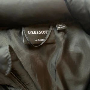 Svart dunväst från Lyle & Scott - Snygg svart dunväst från Lyle & Scott med gul logga på bröstet. Västen har quiltad design, hög krage och dragkedja framtill. Perfekt att slänga över en hoodie eller t-shirt för en chill look.