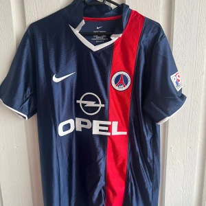 PSG Ronaldinho 21 Nike fotbollströja - Klassisk Paris Saint-Germain fotbollströja med Ronaldinho 21 på ryggen. Tröjan är marinblå med en röd vertikal rand, vita detaljer och PSG-emblem på bröstet. Tillverkad i lätt och ventilerande material, perfekt för fotbollsälskare.