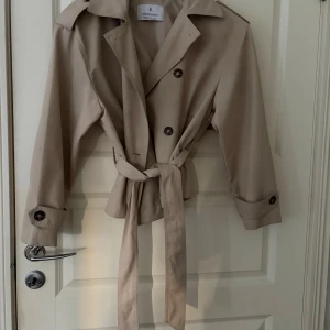 Beige trenchcoat från Stradivarius - Stilren beige trenchcoat från Stradivarius med klassisk krage, knappar och bälte i midjan. Jackan har långa ärmar med knappdetaljer vid ärmslut och en snygg, tidlös look. Perfekt för dig som gillar minimalistisk stil. Diskuterar pris vid snabbaffär! 
