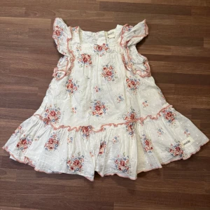 Blommig klänning - Supersöt vit klänning från Newbie med blommigt mönster och två lager. Storlek 62 (2-4mån). Helt oanvänd, endast upptvättad. Knappar i ryggen.