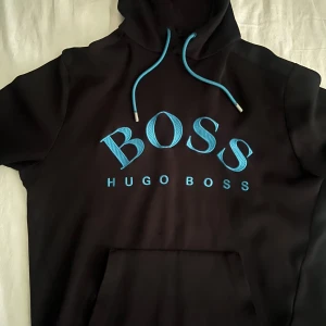 Svart hoodie från Hugo Boss med blå logga - Svart hoodie från Hugo Boss med stor blå BOSS-logga och matchande blå snören i huvan. Klassisk känguruficka framtill och mjukt material som känns skönt mot huden. Perfekt för dig som gillar stilrena streetwear-plagg med tydlig branding.