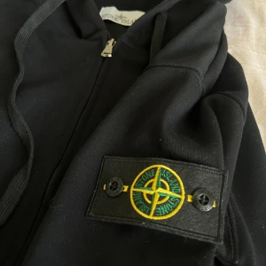 Svart hoodie från Stone Island - Svart hoodie från Stone Island med dragkedja framtill, huva med snörning och klassisk logotyp-patch på ärmen. Tröjan har känguruficka och ribbade muddar. Tillverkad i mjuk bomull, perfekt för en chill och stilren look.