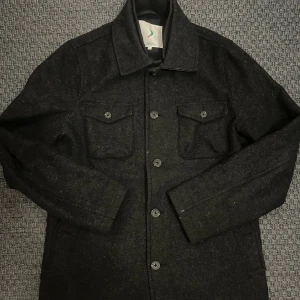 Mörkgrå overshirt från Boomerang - Stilren mörkgrå overshirt från Boomerang i ullblandning. Jackan har klassisk krage, två bröstfickor med lock och knappar framtill med Boomerang-logga. Perfekt lager-på-lager-plagg för kyligare dagar.