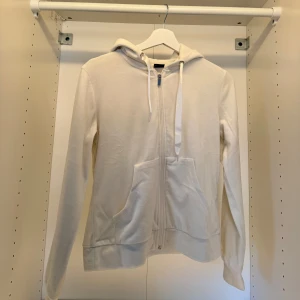 Vit hoodie med dragkedja och huva - Vit hoodie med dragkedja framtill, huva med snörning och två stora fickor. Tröjan har lång ärm och är tillverkad i mjukt bomullsmaterial. Perfekt för en chill och avslappnad stil.