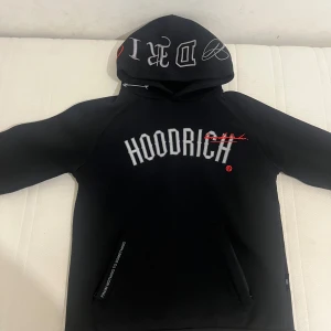 Svart hoodie från Hoodrich med tryck - Priset kan diskuteras!!!Svart hoodie från Hoodrich med stor reflekterande logga på bröstet, broderade detaljer på huvan och tryck på ryggen. Klassisk känguruficka framtill och textdetaljer på fickan. Perfekt streetstyle-plagg med snygga kontraster och cool design.