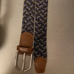 Blått och grått flätat bälte - Snyggt flätat bälte i blått, grått och svart med detaljer i brunt skinn. Bältet är 1 meter. ( pris kan diskuteras) 